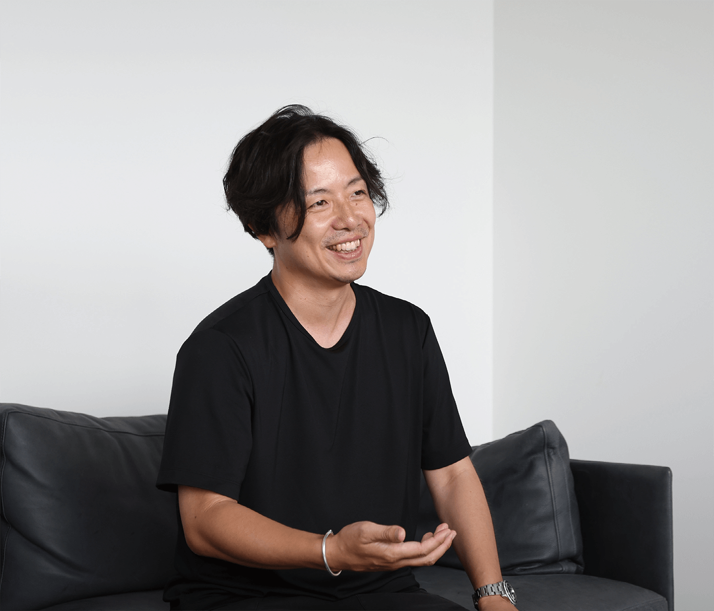 黒いTシャツを着た黒髪のミディアムヘアの男が、明るくミニマルな部屋の暗いソファに座りながら微笑んでいる。.
