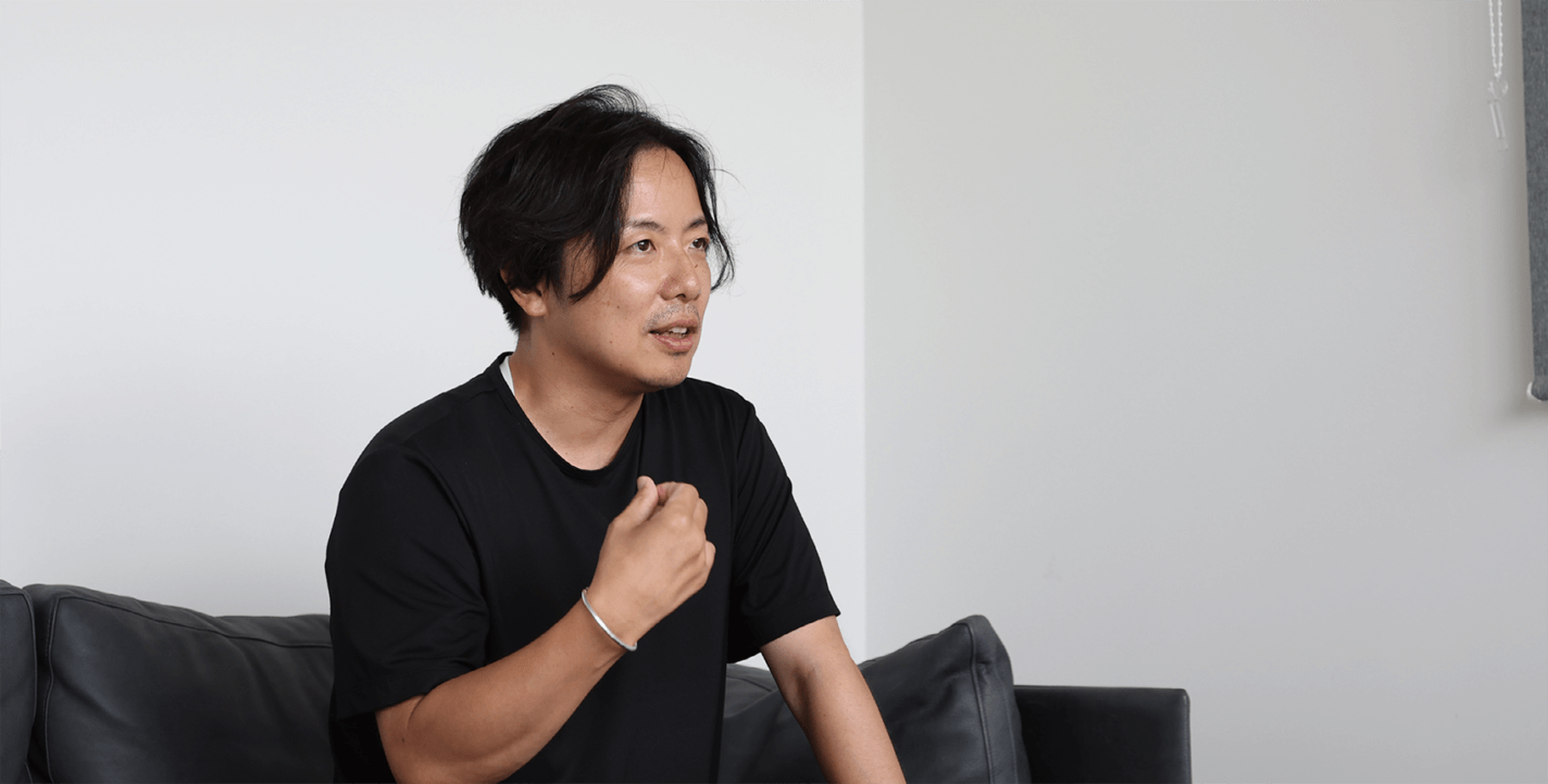 黒いTシャツを着た黒髪のミディアムヘアの人物が、黒っぽいソファに座り、右手でジェスチャーをしながら話している。背景は真っ白な壁。.