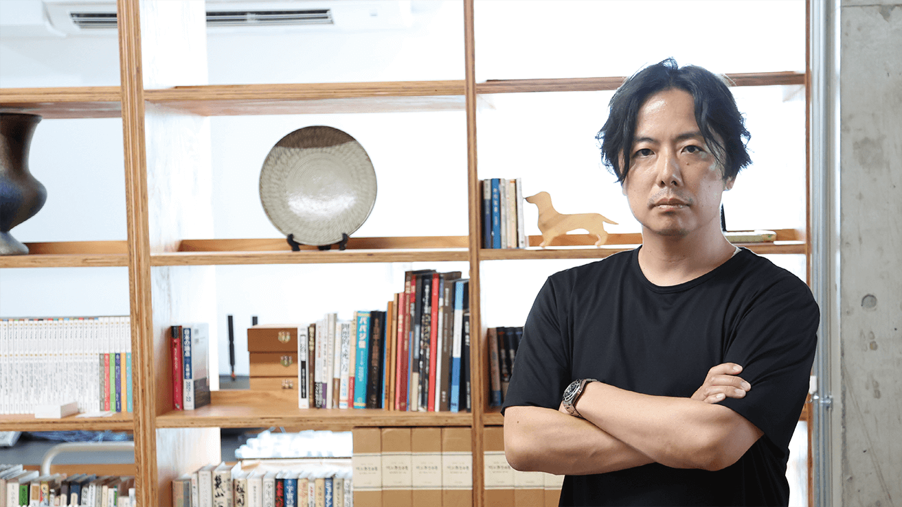 モダンで明るい部屋の、本や装飾品でいっぱいの木製の本棚の前に、黒いTシャツを着た黒髪の男が腕組みをして立っている。.