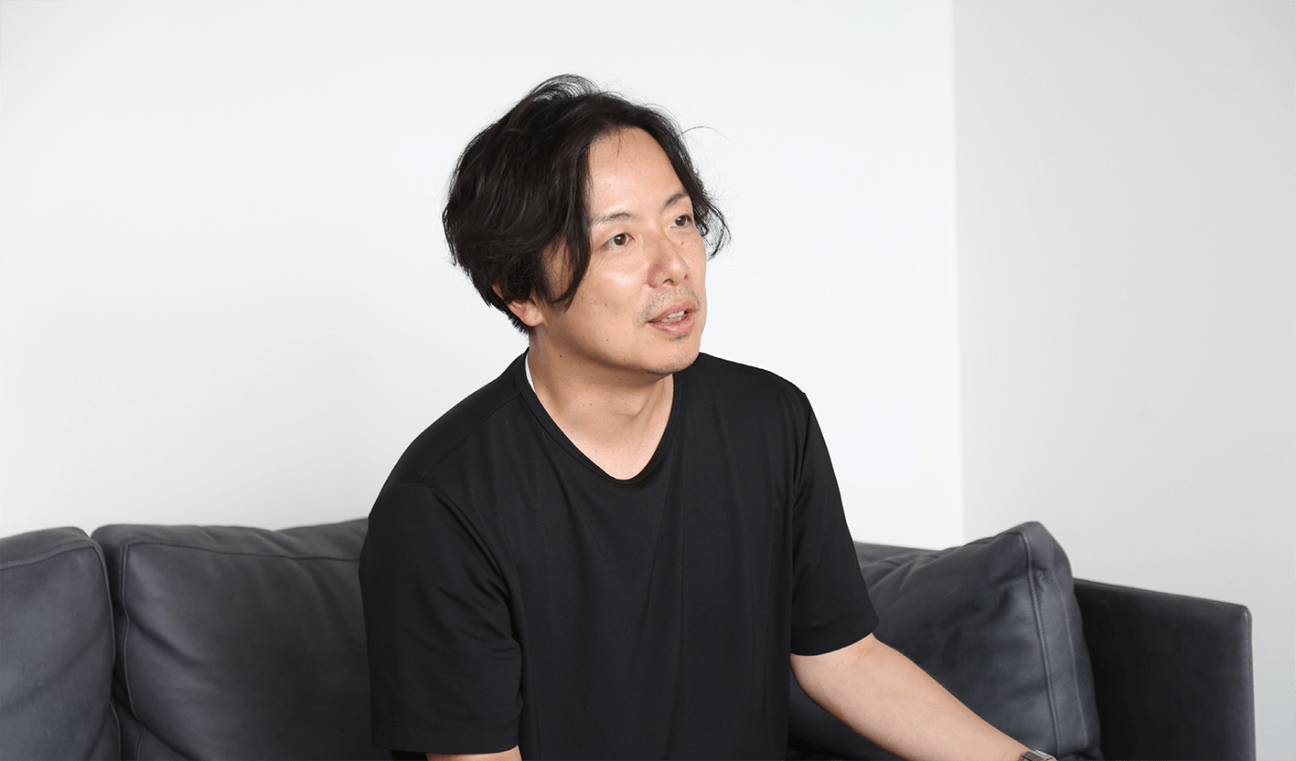 黒いTシャツを着た黒髪のミディアムヘアの男が、真っ白な壁を背に灰色のソファに座り、やや横を向いて思案顔をしている。.