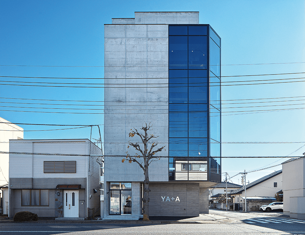 コンクリートのファサードに大きなガラス張りの角を持つ4階建ての近代的なビルが、赤いドアの小さな白い家の横に建っている。澄み切った青空の下、葉のない木が建物の前にある。.