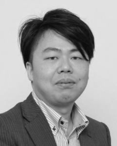 ピンストライプのスーツジャケットにストライプの襟付きシャツを着た黒髪の短髪の男性が、無地の背景の中、ニュートラルな表情でカメラに向かっている。.