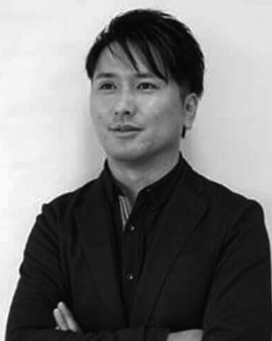 スーツの上着と襟付きシャツを着用し、腕を組んでやや横を向いて立っている黒髪短髪のアジア人男性の白黒写真。.