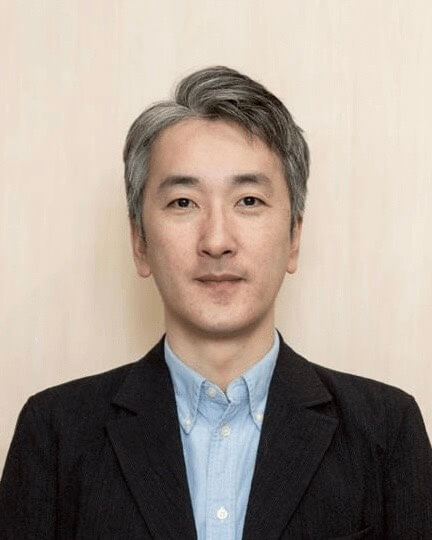 白髪と黒髪のショートヘアの男性が、水色のボタンアップシャツの上にダークブレザーを着ている。カメラに向かい、わずかに微笑み、背景はニュートラルなベージュ。.