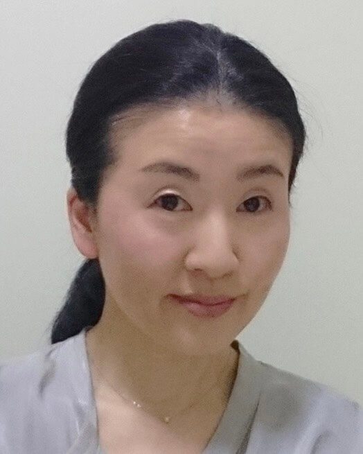 長い黒髪を後ろで束ね、薄いグレーのトップスを着た女性が、無地の明るい背景の中、優しく微笑みながらカメラを見つめているポートレート。.