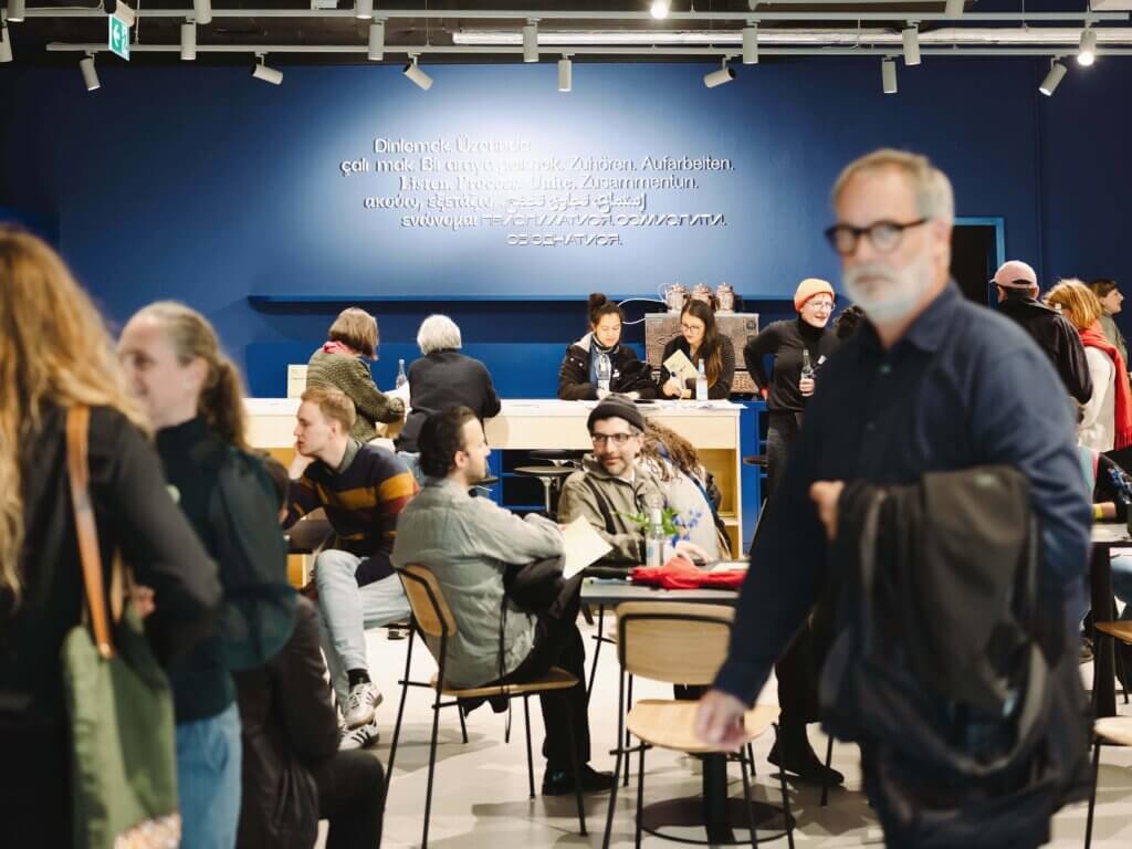 Ein belebtes modernes Café mit Menschen, die an Tischen sitzen, sich unterhalten, lesen und arbeiten. Ein Mann mit Brille und dunkler Jacke geht im Vordergrund vorbei. Im Hintergrund sind blaue Wände mit weißem, mehrsprachigem Text zu sehen.
