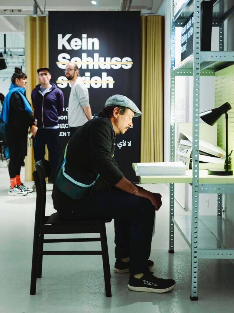 Ein Mann sitzt an einem Schreibtisch und liest ein Buch in einem modernen Galerieraum. Hinter ihm stehen drei Personen neben einem schwarzen Schild mit weißer und gelber deutscher Schrift: Kein Schlussstrich. Regale und eine Lampe sind zu sehen.