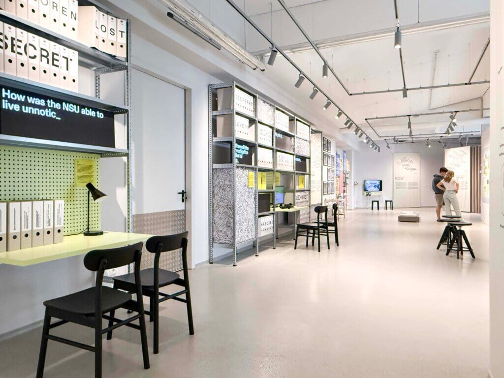 Ein moderner, minimalistischer Ausstellungsraum mit heller Beleuchtung, schwarzen Stühlen, Schreibtischen und Regalen mit beschrifteten Ordnern. Zwei Personen betrachten die Displays an der gegenüberliegenden Wand. Texttafeln und Dokumente sind im ganzen Raum zu sehen.