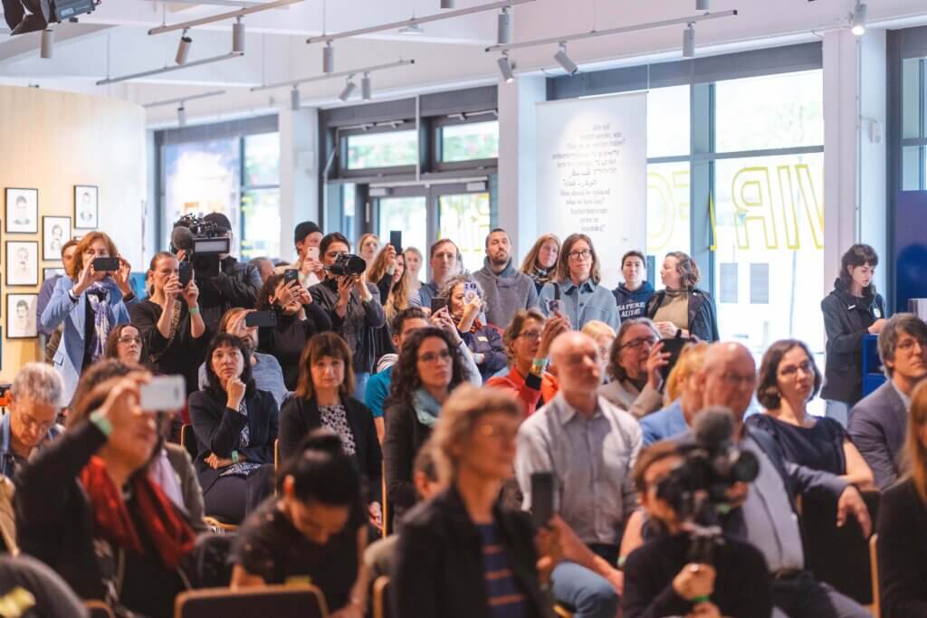 Eine bunt gemischte Gruppe von Menschen sitzt und steht in einem hellen Raum, viele halten Telefone oder Kameras in der Hand und beobachten aufmerksam eine Veranstaltung oder Präsentation.