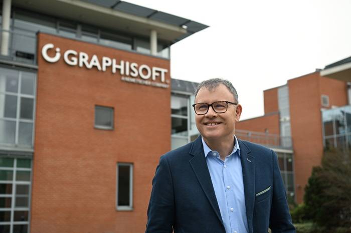 Ein Mann im Anzug und mit Brille steht lächelnd vor einem modernen Backsteingebäude mit der Aufschrift GRAPHISOFT. Der Himmel ist bedeckt.
